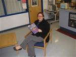 Mrs. Quinn sitting in a chair reading a book and smiling.